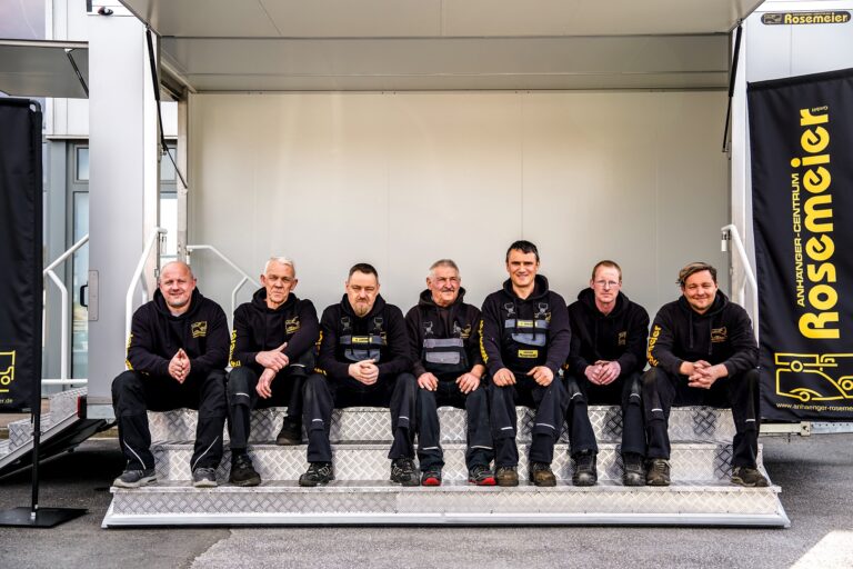 Sieben Männer in schwarzer Arbeitskleidung sitzen nebeneinander auf Metallstufen vor einem Firmengebäude. Links und rechts sind schwarze Banner mit gelbem 'Rosemeier'-Logo zu sehen.